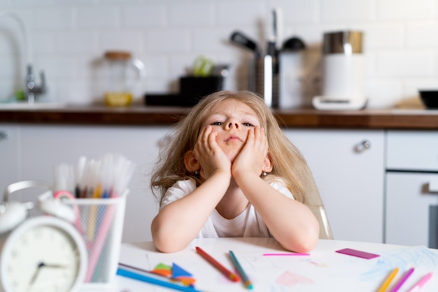 Tired little blonde girl at home at the kitchen table, the concept of home education.