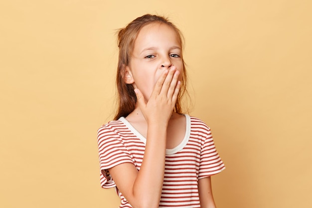 Tired little brown haired girl wearing striped tshirt standing isolated over beige background covering mouth with palm yawning felling sleepy