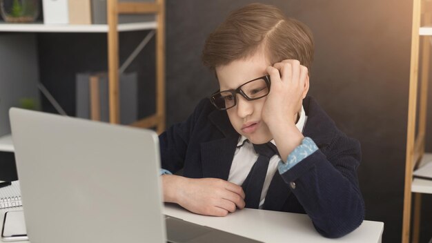 Piccolo imprenditore stanco che lavora al computer portatile in ufficio. ragazzo con gli occhiali e abito formale che finge di essere un grosso impiegato