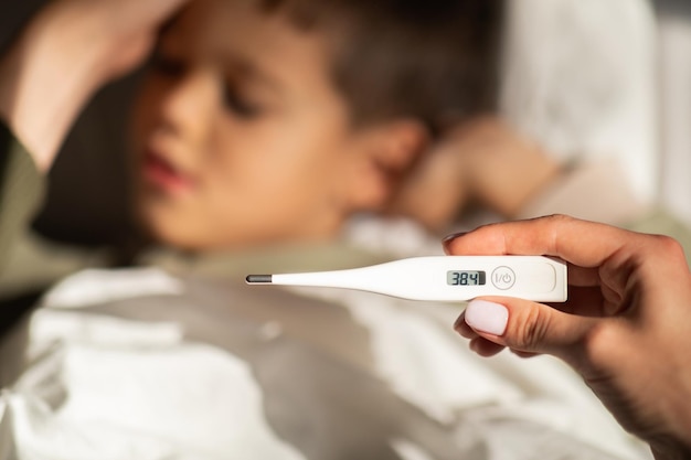 tired little european boy lies on bed sleeping mom measures temperature with thermometer in bedroom