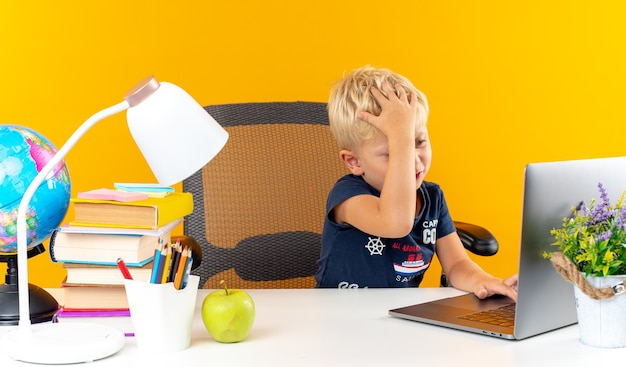 Tired little school boy sitting at table with school tools used laptop putting hand on head 
