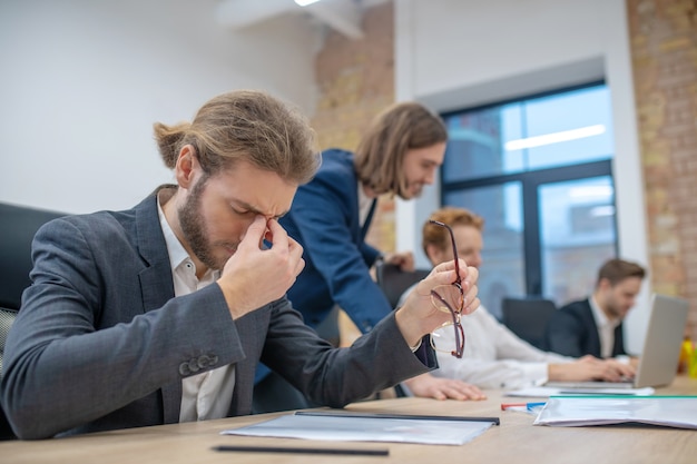 Tired longhaired young man closing eyes and taking off glasses sitting in office over document and colleagues