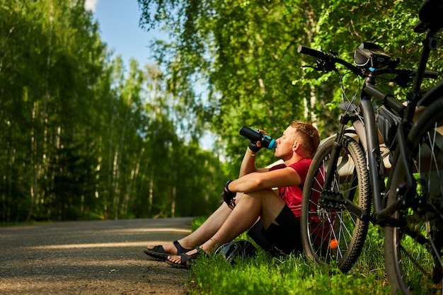tired male cyclist sits on the side of the road and drinks water from a bottle.