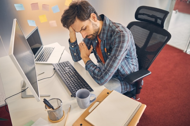 tired male worker looking down and propping up head with hand while sitting at the table with computer and laptop