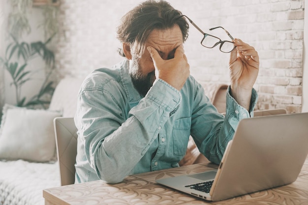 写真 疲れた男がガラスを持って触れているのは、ノートパソコンで仕事をした後の目 オンライン活動と解決すべき問題のコンセプト テーブルデスクでラップトップを使用する現代の労働者 経済的困難