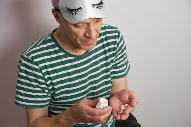 Tired man in a sleep mask pours out insomnia pills from a jar on a white background insomnia of the elderly