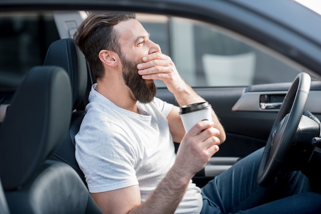 行くためにコーヒーを持って車の前部座席であくびをしている疲れた男