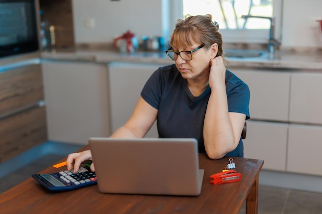 Tired mature blond woman freelancer working from home using laptop sitting in kitchen