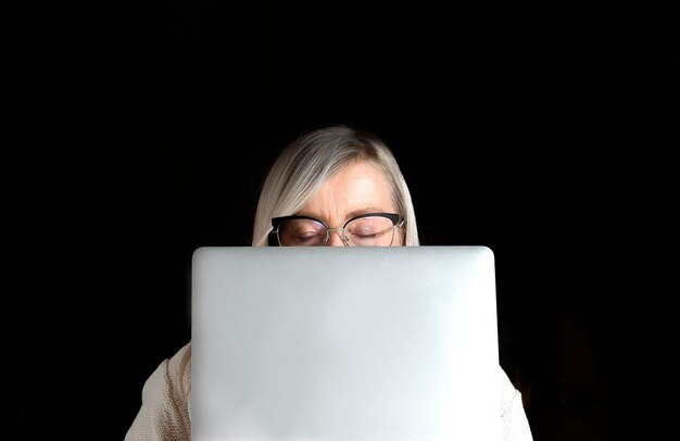 Photo tired mature womans face with closed eyes and glasses hidden behind a computer laptop