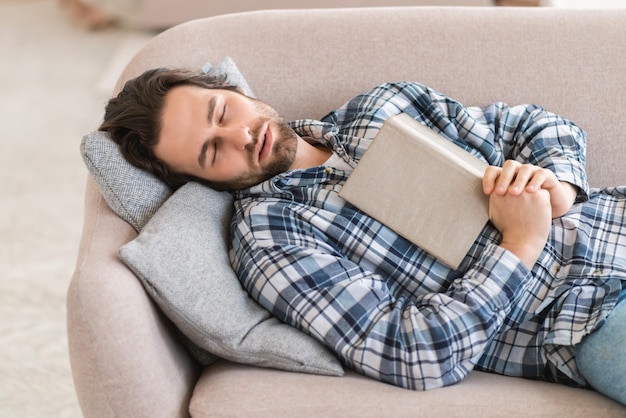 Tired millennial european man in domestic clothes with stubble sleeps with book diary lies on sofa in living room