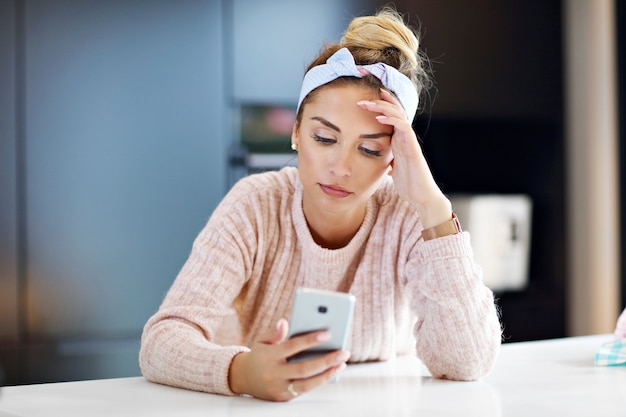 Tired millennial woman resting head in the kitchen and using smartphone