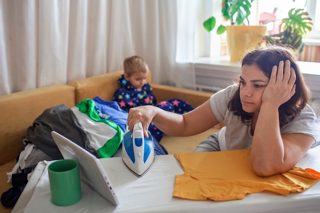 Photo tired mom watches video on tablet and ironing things next to her son with smartphone too