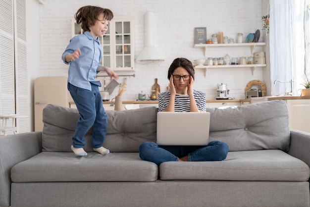 Tired mother sit on couch work on laptop at home hyperactive little kid jumping drawing attention