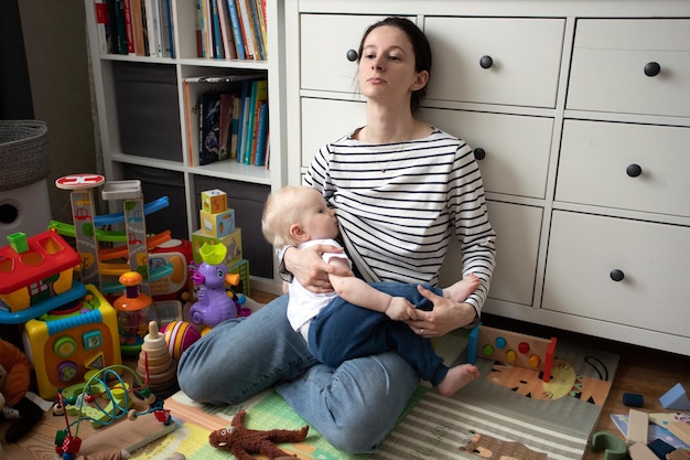 уставшая мать с малышом на руках сидит в куче игрушек в беспорядке и кормит грудью