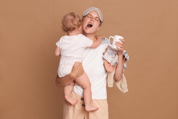 Tired mother yawning and feels tired holding baby stuff on her shoulder standing indoor isolated over brown wall holding cup of coffee and keeps eyes closed