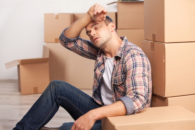 Tired of moving boxes. Tired young man sitting on the floor and leaning at the carton box stack while more cardboard boxes laying on the background