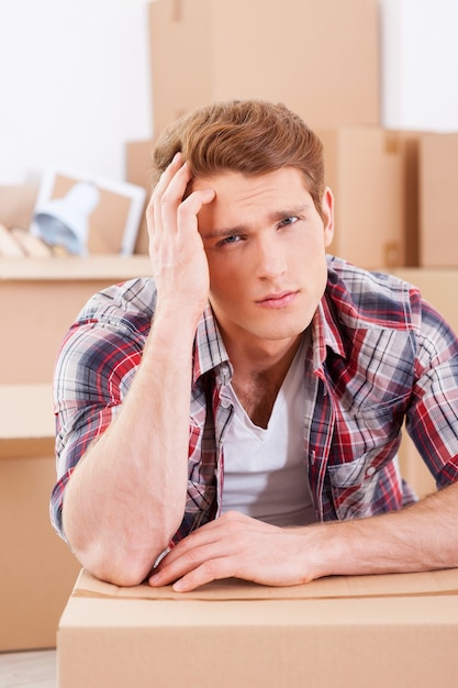 Tired of moving. Depressed young man sitting on the floor and holding hand in hair while cardboard boxes laying on the background