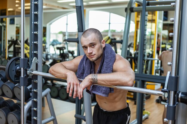 Tired muscular man with a naked torso and a towel on his neck resting on a barbell in the gym