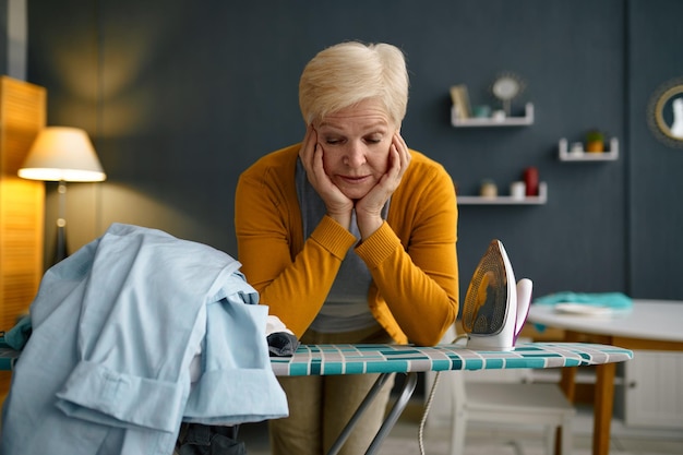 Tired old woman standing front of ironing board with clothes pile. Fatigue from household chores