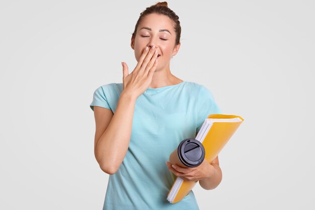 Tired overworked comely student has sleepy expression, yawns as needs rest, covers mouth with palm, holds takeaway coffee and folder, isolated over white. People and deadline concept