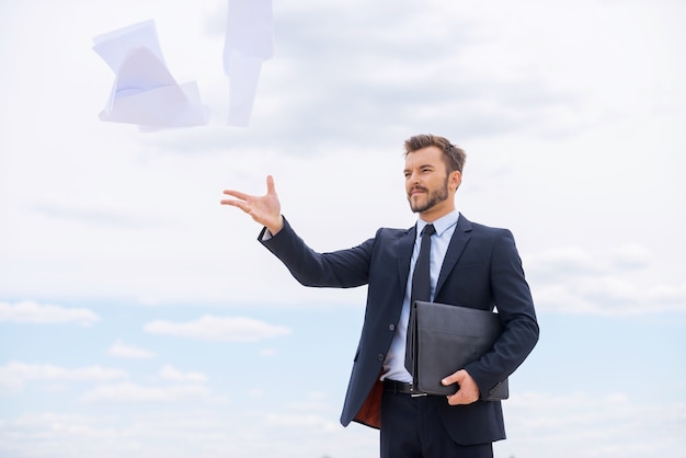 Tired of paperwork. Frustrated young businessman throwing documents away while standing against blue sky