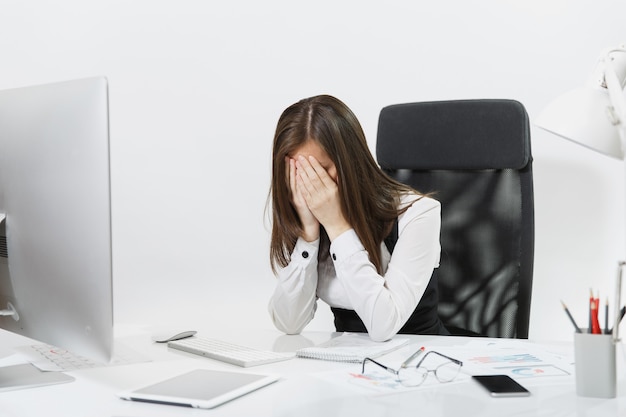 Stanco perplesso e stressato donna d'affari dai capelli castani in abito seduto alla scrivania, coprendosi il viso con le mani, lavorando al computer contemporaneo con documenti in ufficio leggero