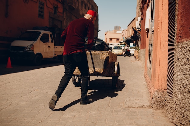 Tired porter man with heavy trolley and tourist luggage walking at street to hotel in Marrakesh, Morocco.