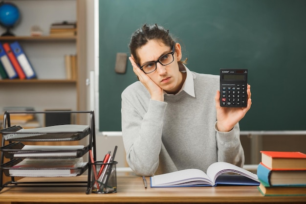 tired putting hand on cheek young male teacher holding calculator sitting at desk with school tools on in classroom