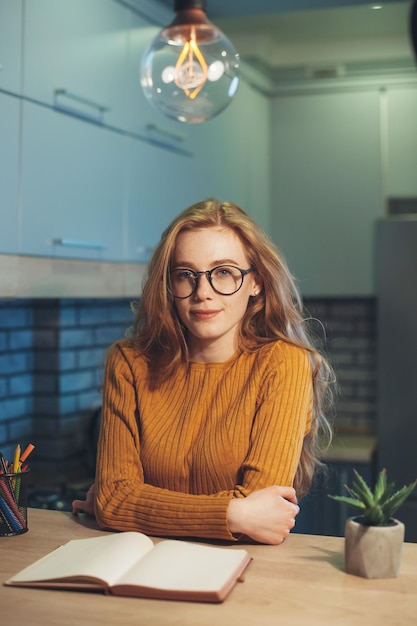 Tired redheaded woman student sitting at a table with textbooks looking at camera preparing for coll