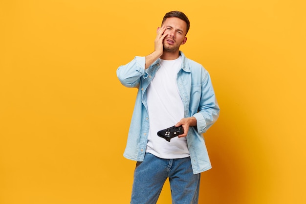 Tired sad tanned handsome man in blue basic tshirt lose game\
hold joystick gamepad touches cheek posing isolated on orange\
yellow studio background copy space banner mockup people lifestyle\
concept