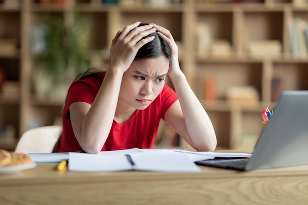 Tired sad teenage asian girl study find mistake hold her head with hands looks at pc in room