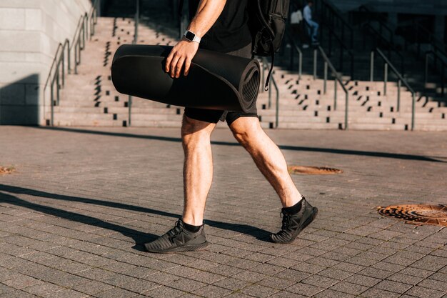 Tired and satisfied male sportsman returns home after morning\
workout. cut view of sporty man\'s legs. urban sport concept. man\
walking on street and holding rolled yoga mat.