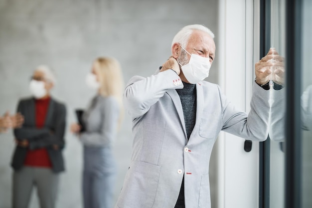 코로나 바이러스나 COVID-19 전염병 동안 현대 사무실에 서서 창 밖을 내다보고 있는 보호 마스크를 쓴 피곤한 고위 사업가.