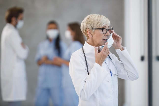 A tired senior doctor holding protective mask and pensive looking out the window while standing in a hospital hallway during the Covid-19 pandemic.