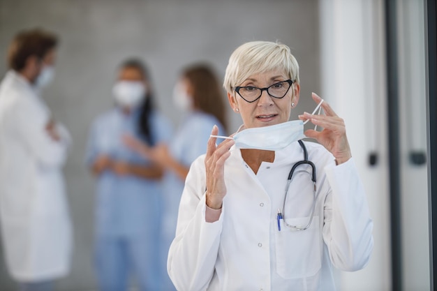 A tired senior doctor standing and holding protective mask while having quick break in a hospital hallway during the Covid-19 pandemic. Looking at camera.