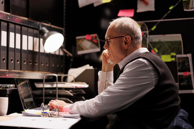 Foto investigatore anziano stanco che pensa alla soluzione per catturare il sospetto, lavorando a tarda notte in un procedimento penale in una stanza buia. ufficiale di polizia oberato di lavoro che controlla le prove della scena del crimine, esaminando il rapporto della vittima