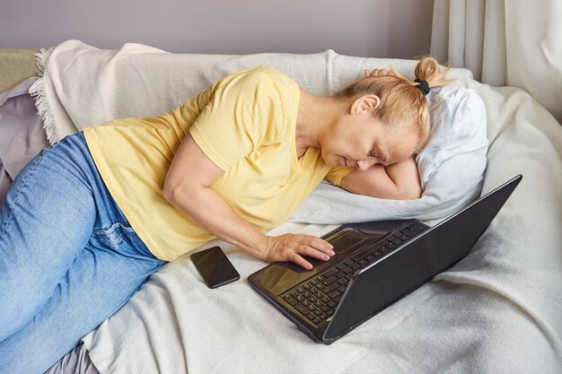 La donna anziana stanca in camicia gialla si è addormentata dopo aver lavorato con il computer portatile sul divano di casa.