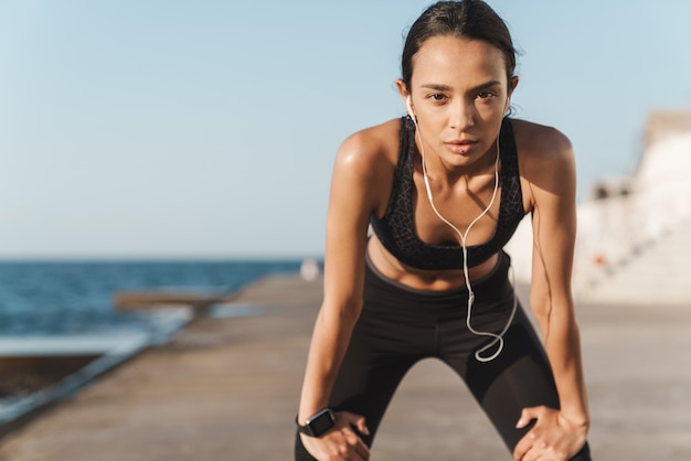  tired serious beautiful young strong sports woman outdoors at the beach at morning have a rest listening music with earphones.