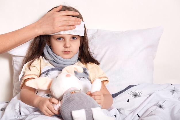 Tired sick little girl having unknown hand on forehead holding wet cold napkin