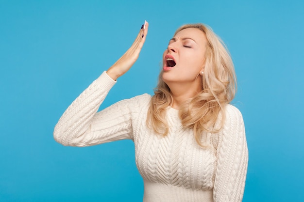 Tired sleepy blond woman in white knitted sweater yawning, closing mouth with hand, exhausted with insomnia, lack of energy. Indoor studio shot isolated on blue background