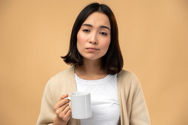 Tired sleepy woman holding cup of coffee, looking at the camera with tired expression, cannot wake up in the morning and go to work. Isolated beige background