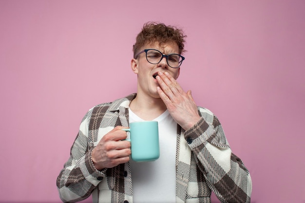 Tired sleepy young guy in glasses holds mug of coffee and yawns on pink background