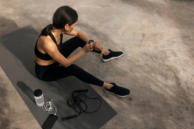 Tired slender caucasian millennial lady in sportswear sits on mat with skipping rope and bottle of water checks pulse