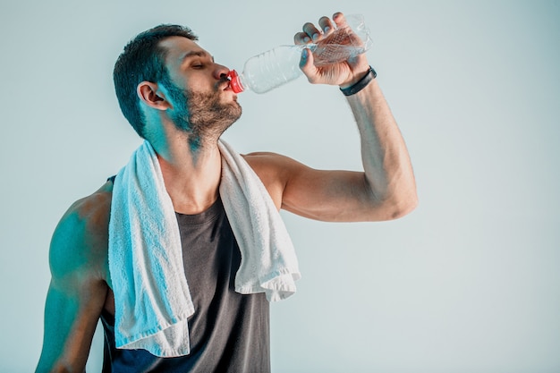 Tired sportsman drinking water from bottle after training. Young bearded european man with closed eyes and towel. Person wear sports uniform. Isolated on turquoise background. Studio shoot