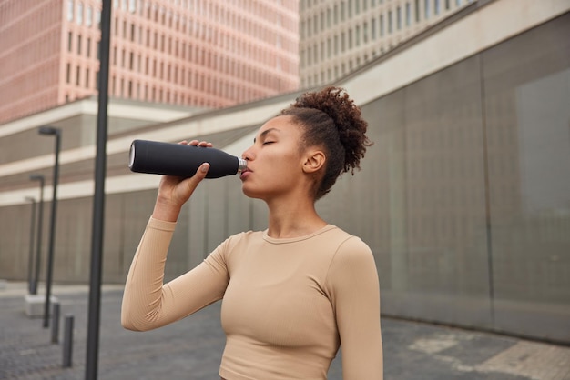 Tired sporty woman with curly combed hair drinks fresh cold water from bottle rests after active jog or intense physical training dressed in sportswear poses at urban setting Recreation time