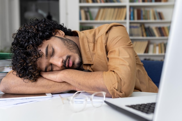 Tired student sleeping on table inside university academic library hispanic man among books with