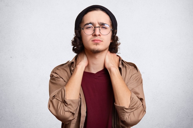 Tired teenager with crisp hair, has pain in neck, dressed in red casual t shirt, brown shirt and black cap