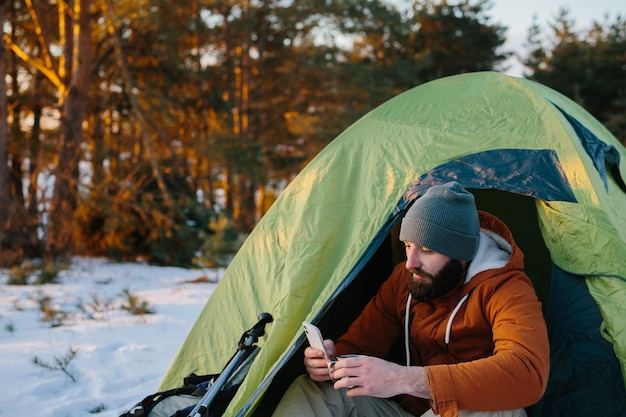 A tired tourist rests after a difficult climb sitting in a tent in a snowy forest A man uses his phone to send a message or check his GPS