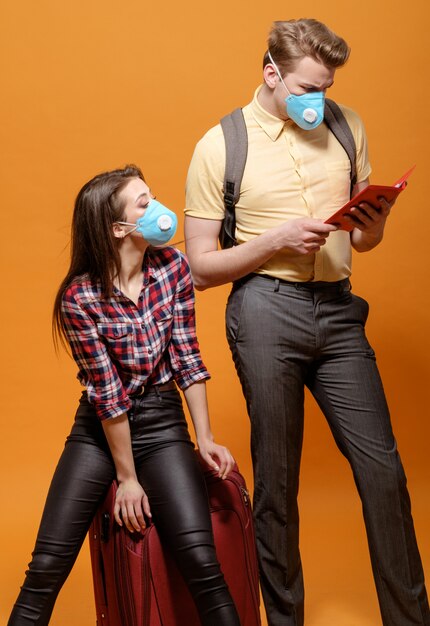 Tired tourists, travelers man and woman in medical masks  on a yellow orange background, coronavirus pandemic, closed border countries,  big suitcase