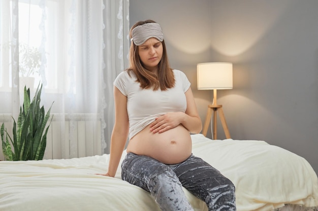 Foto donna incinta stanca e infelice che si tiene la schiena, che sente il disagio, che è esausta, che sente la stanchezza negli ultimi mesi della gravidanza, mentre è seduta a letto a casa.